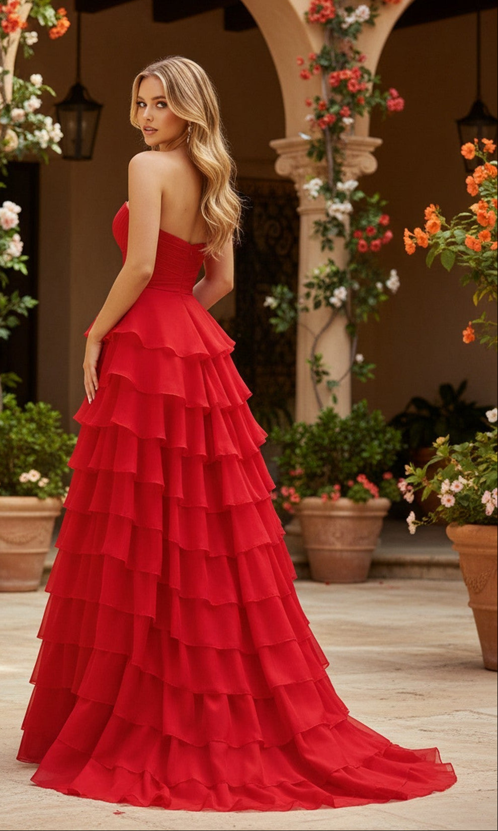 A woman wearing the PromGirl Exclusive 250233B long prom dress with a strapless bodice and tiered ruffle skirt stands on an outdoor tiled patio surrounded by potted plants and flowering vines.